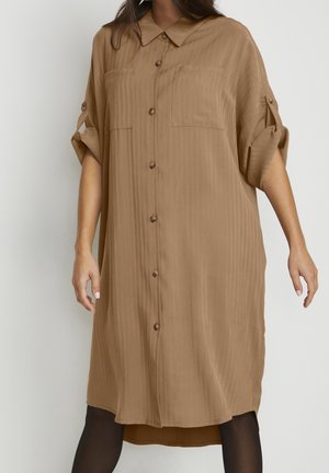 Femme portant une robe-chemise ample couleur brun clair, boutonnée, avec les manches retroussées et deux poches poitrine, debout devant un fond uni.