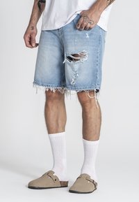 Traper šorts u svijetloplavoj boji s oštećenjima, neurednim rubom i opuštenim krojem. Upareni s bijelim čarapama i bež sandalama otvorenih prstiju.