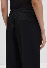 Persoon in zwarte elastische taille broek met een enkele paspel achterzak, getoond van taille tot halverwege de dij, staand tegen een effen achtergrond.