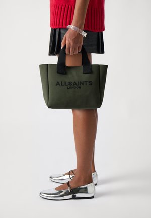 Zöld vászontáska fekete fogantyúkkal és hímzett "ALLSAINTS LONDON" logóval, rózsaszín bordázott pulóverrel és ezüst balerina cipőkkel párosítva.
