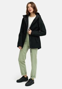 Femme portant une veste à capuche noire, une chemise blanche, un pantalon vert clair retroussé et desSneakers noires, se tenant devant un fond uni.