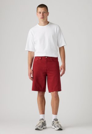 Ung mann iført en ensfarget hvit t-skjorte, røde knelange shorts, hvite sokker og grå løpesko står mot en nøytral bakgrunn.