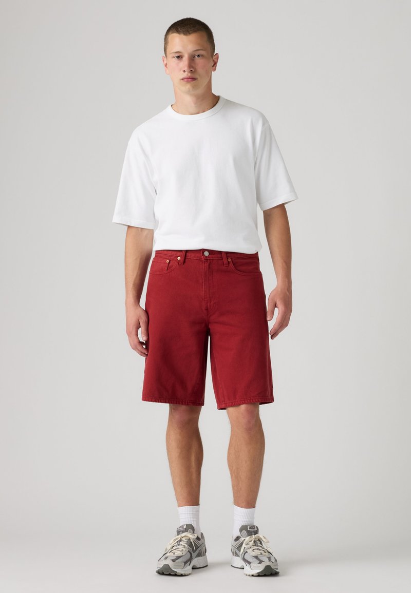 Joven con una camiseta blanca sencilla, pantalones cortos rojos hasta la rodilla, calcetines blancos y zapatillas de correr grises, de pie frente a un fondo neutro.