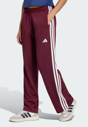Person iført bordeaux Adidas træningsbukser med hvide striber og Adidas-logo, kombineret med hvide sneakers med sorte striber.