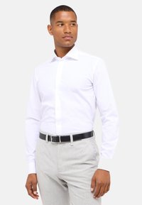 Witte lange mouw dresseshirt met een puntige kraag en knoopsluiting, gecombineerd met lichtgrijze textuurbroek en een zwarte riem.