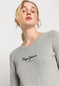 Grå langermet topp laget av bomullsblanding, med rund hals og svart "Pepe Jeans London" logo på brystet. Glatt tekstur.