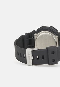 Zegarek G-SHOCK z czarnym gumowym paskiem, srebrną metalową klamrą i teksturowaną obudową. Posiada stalowy tył z wygrawerowanymi detalami.