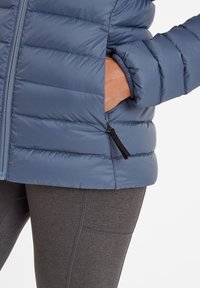 Blue puffy jacket with quilted design and elastic cuffs, paired with gray leggings, featuring a side pocket with a black zipper pull.