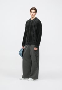 Crni pleteni kardigan s uzorkom, zatvaranjem na gumbe i ovratnikom, uparen s širokim sivim hlačama s tankim prugama i bočnim vezom.