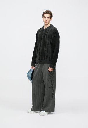 Melna adīta kardigāna ar rakstu, pogu aizdari un apkaklīti, kombinēta ar plašām pelēkām svītrainām biksēm ar sānu izšuvumu.