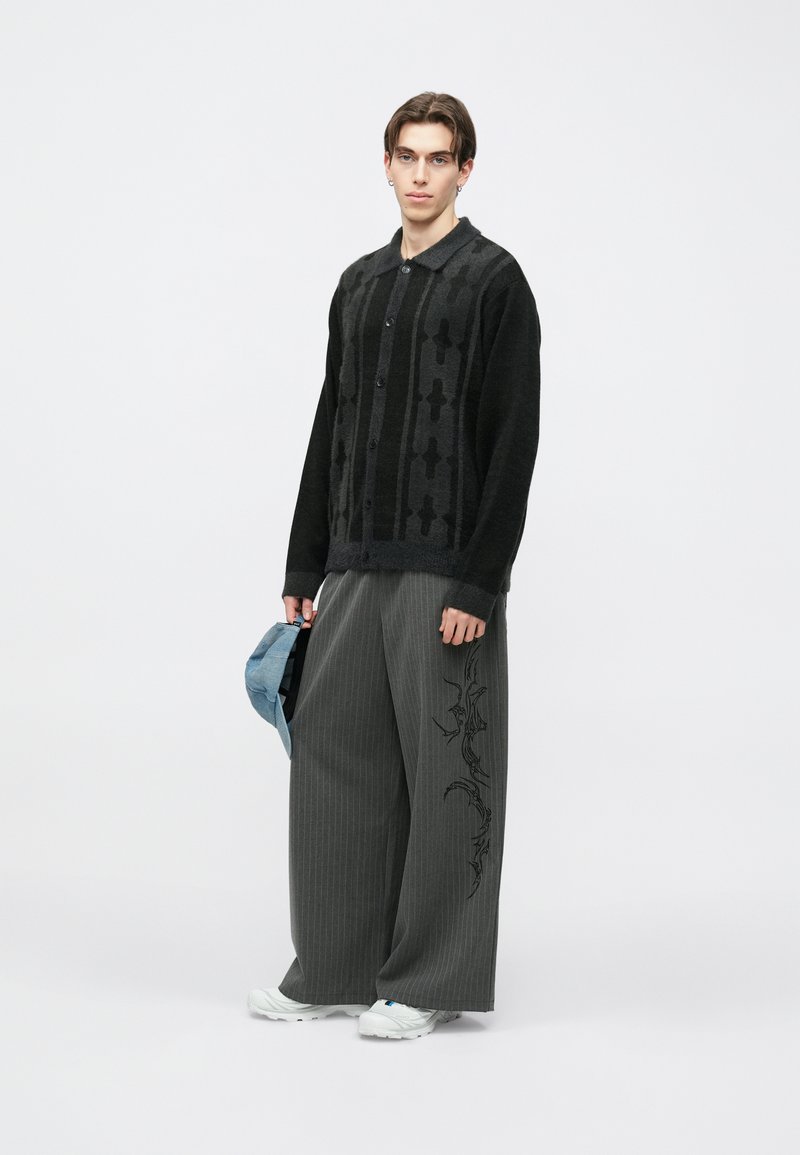 Crni pleteni kardigan s uzorkom, zatvaranjem na gumbe i ovratnikom, uparen s širokim sivim hlačama s tankim prugama i bočnim vezom.