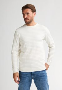 Man met kort haar en baard, gekleed in een textuur witte lange mouwen trui en blauwe spijkerbroek, staand met één hand in zijn zak.