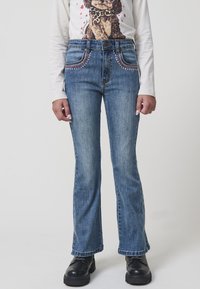 Jeans acampanados de denim azul con costuras decorativas en los bolsillos frontales. Presenta un cierre de botón y un ajuste recto, con una textura ligeramente desgastada.