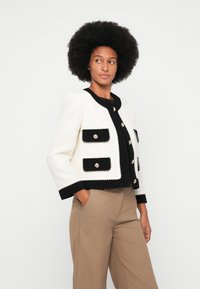 Veste en tweed crème et noir avec quatre poches, boutons dorés et bordure texturée. Portée par-dessus un haut noir et un pantalon beige.