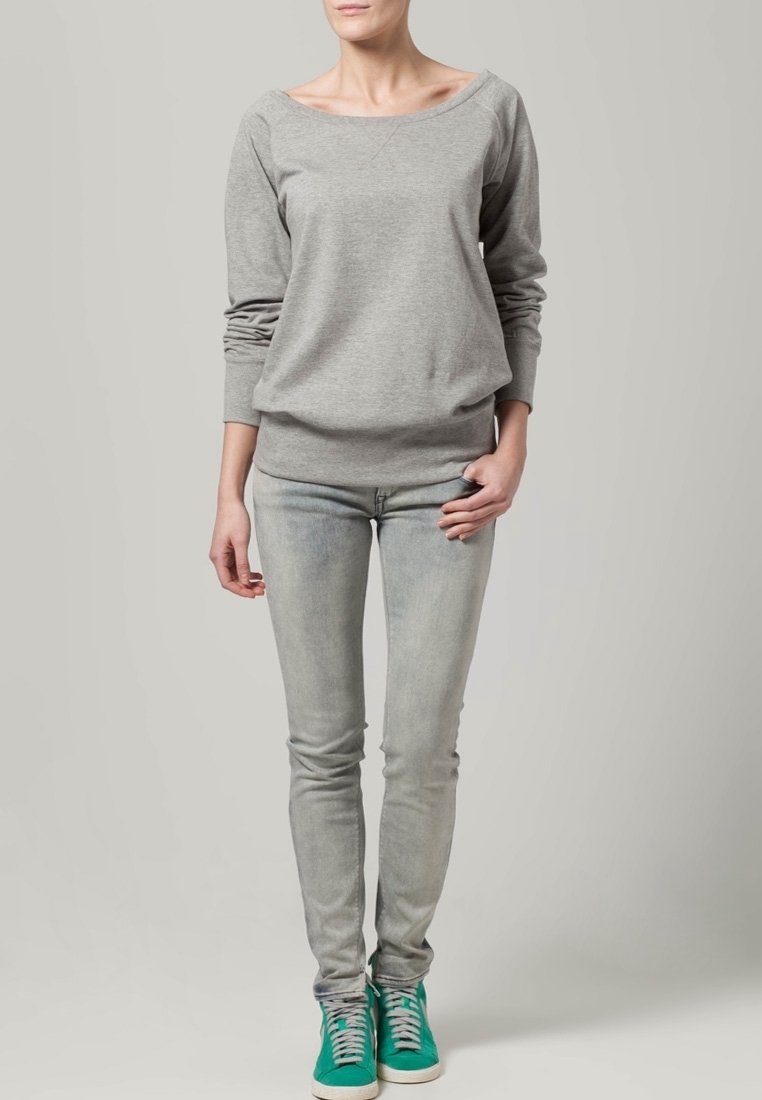Femme portant un haut à manches longues gris clair, un jean skinny délavé et des baskets vertes, debout devant un fond gris clair uni.