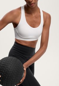 Weißer Sport-BH mit rundem Halsausschnitt, kombiniert mit schwarzen, hoch taillierten Leggings. Die Person hält einen strukturierten schwarzen Gymnastikball.