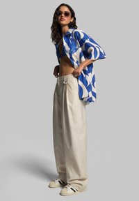 Short-sleeve shirt in blue and white abstract pattern, paired with cream wide-leg pants and beige sneakers with black stripes, worn by a model.