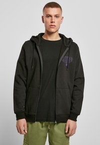 Sudadera negra con cremallera y capucha con cordón, bolsillos delanteros y un sutil detalle de logo púrpura. Combinada con una camiseta negra y pantalones verdes.