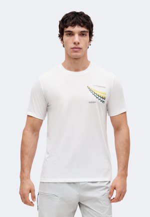 Mladý muž, ktorý má na sebe biele tričko s okrúhlym výstrihom a žlto-čiernym geometrickým vzorom na ľavej strane hrudníka, v kombinácii so svetlošedými nohavicami.