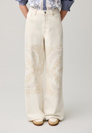 Pantalones blancos de talle alto y pierna ancha con patrones florales intrincados en beige, combinados con zapatos beige sin cordones y una camisa de rayas metida por dentro.