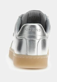 Sneaker metallico argento con una texture liscia, suola beige chiaro e logo "GUESS" in rilievo sul tallone. Design casual, vista posteriore.