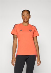 Korte mouwen, koralen sportshirt met blauwe accenten, ronde hals en logo-detail. Materiaal lijkt lichtgewicht en ademend te zijn.
