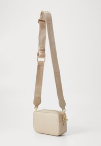 Sac en cuir beige, de forme rectangulaire, porté en bandoulière. Il est doté d'une fermeture éclair dorée et d'une large bandoulière embossée avec "VALENTINO". Texture lisse.