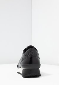 Baskets noires avec panneau de talon texturé, dessus noir, semelle intermédiaire blanche et semelle extérieure noire épaisse, vues de l'arrière sur une surface blanche.