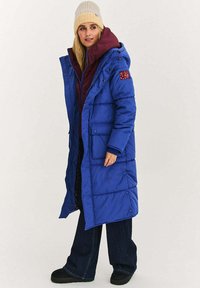 Niebieski pikowany długi płaszcz z kapturem, z bordo podszewką, dwoma przednimi kieszeniami oraz teksturowaną beżową dzianinową czapką. Całość dopełniają ciemne spodnie.