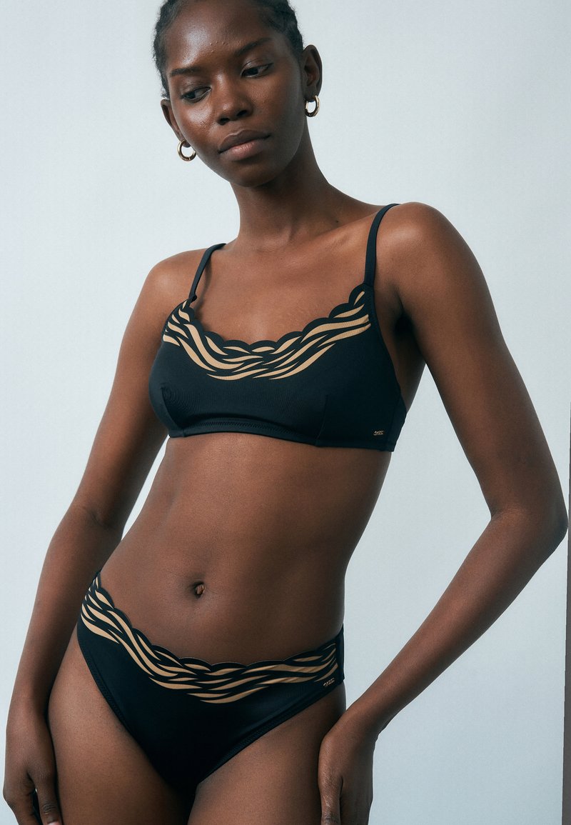 Γυναίκα που φοράει μαύρο σετ εσώρουχα με μπεζ κυματιστό μοτίβο, χρυσούς κρίκους σκουλαρίκια, στέκεται μπροστά σε απλό ανοιχτό γκρι φόντο.