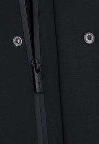 Czarna kurtka z materiału o gładkiej teksturze, z centralnym zamkiem błyskawicznym z logo oraz metalowym zatrzaskiem jako akcent.