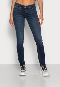 Jeans skinny azul oscuro con cierre de botón y cremallera, diseño de cinco bolsillos y sutiles detalles deslavados. Combinados con zapatillas blancas y negras.