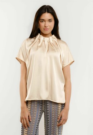 Young woman with long dark hair wearing a shiny cream blouse with pearl buttons and patterned pants, standing against a plain background.