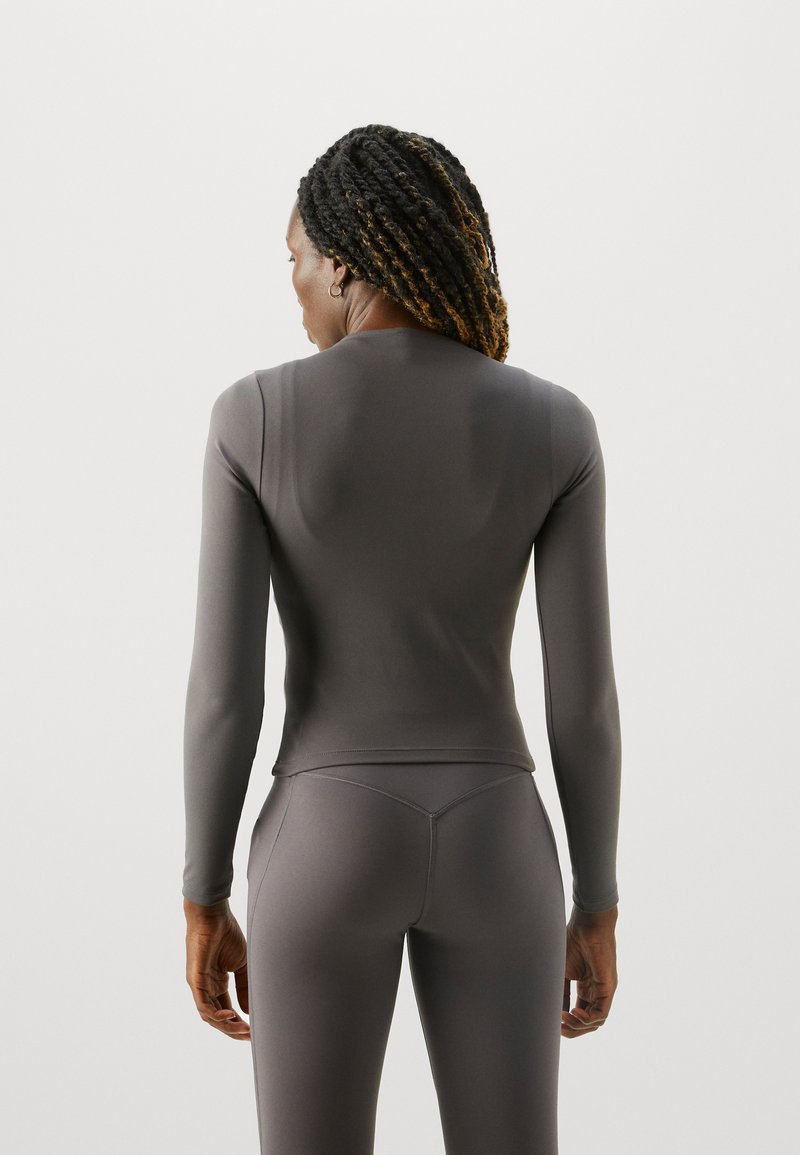Haut ajusté à manches longues en tissu gris foncé, texture lisse, avec un col rond et des coutures subtilement courbées. Associé à des leggings assortis.