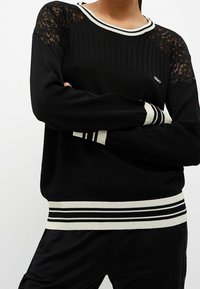 Czarny sweter z prążkowaną teksturą, koronkowymi rękawami oraz kontrastowymi białymi i czarnymi paskami przy dekolcie i u dołu.