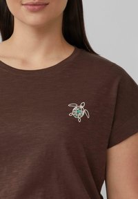 Bruine T-shirt met korte mouwen met een witte geborduurde schildpaddesign en turquoise accenten. De stof lijkt zacht en gestructureerd.