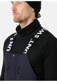 Navy waterdichte overalls met verstelbare witte banden. Voorzien van twee voorzakken met rits en een hoge kraag, gedragen over een zwarte ribbeltop.