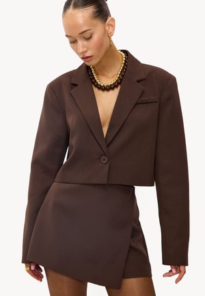 Vrouw draagt een donkerbruine cropped blazer en een asymmetrische minirok met gelaagde bruine en gouden kralenkettingen en gouden oorringen.