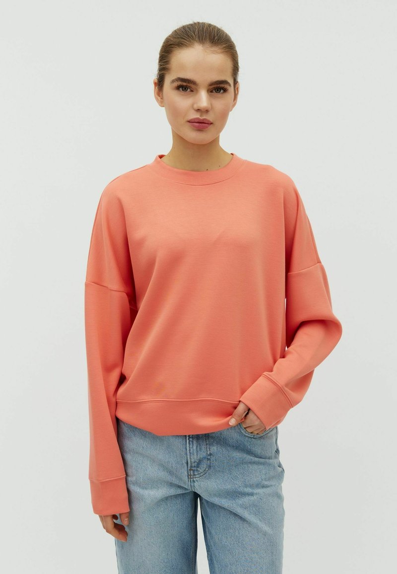 Giovane con capelli tirati indietro che indossa una felpa oversize color corallo e jeans azzurri, in piedi contro uno sfondo bianco semplice.