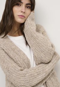 Cardigan beige texturé avec une coupe décontractée et des manches larges, superposé sur un haut blanc, présentant un design en tricot confortable. Teint lisse et cheveux bruns.