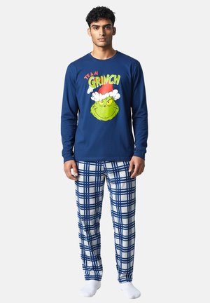 Mann trägt blaues Langarmhemd mit "Team Grinch" und Grinch-Gesicht, blaue und weiße karierte Schlafanzughose und weiße Socken, steht vor einfarbigem Hintergrund.