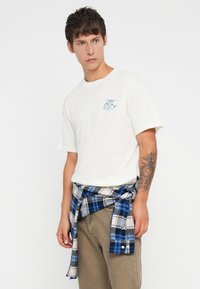 White cotton t-shirt with blue and green embroidered design on the left chest, paired with plaid shirt tied at the waist in blue and black.