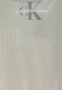 Calvin Klein Jeans taske, lavet af glat, let stof, har et stort gråt logo med en markant tekstureret design på en lys baggrund.