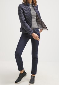 Veste matelassée bleu marine avec coutures en chevrons, fermeture zippée à l'avant et col montant, portée sur un t-shirt à manches longues rayé, associée à un jean foncé et des chaussures noires sans lacets.