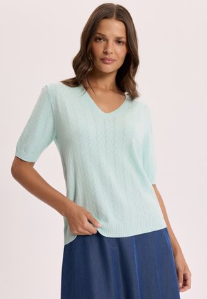 Kobieta ubrana w jasnoniebieski krótki sweter z dekoltem w serek oraz ciemnoniebieską spódnicę, stojąca na tle jasnego, jednolitego tła.