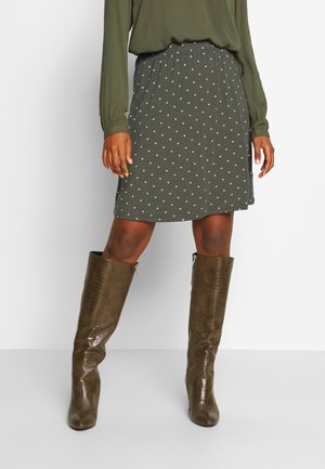 Frau trägt ein olivgrünes langärmliges Oberteil, einen kniehohen dunkelgrünen Rock mit weißen Punkten und hohe braune Lederstiefel mit Struktur.