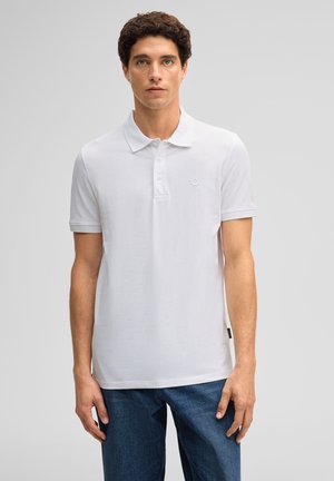 Junger Mann mit lockigem Haar, der ein weißes Poloshirt mit kurzen Ärmeln und blaue Jeans trägt und vor einem schlichten hellgrauen Hintergrund steht.