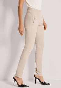 Vrouw in beige slim-fit broek en zwarte gepatineerde hoge hakken, staand tegen een neutrale achtergrond.