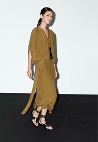 Robe portefeuille verte olive avec des manches chauve-souris, une ceinture à la taille, et des sandales plates noires avec des sangles métalliques, portée par un mannequin sur un fond clair.