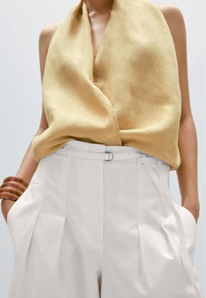 Haut sans manches jaune moutarde rentré dans un pantalon blanc taille haute avec ceinture, mains dans les poches, et bracelets en bois à un bras.